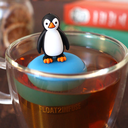 penguin tea infuser for loose leaf tea with a fine mesh stainless steel tea strainer that reads FLOAT2INFUSE on a blue base floating in a glass tea mug with books in the background