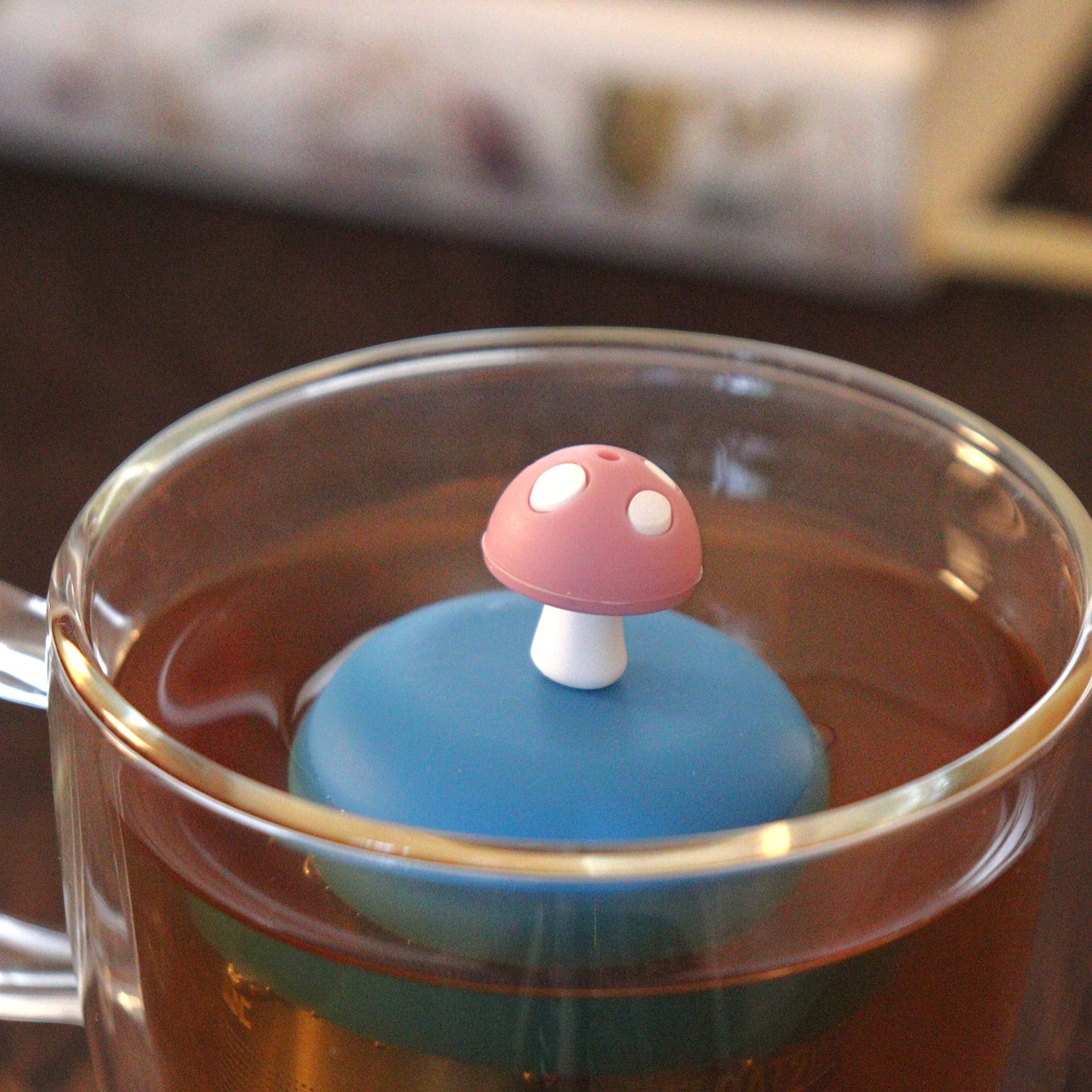 pink mushroom tea infuser for loose leaf tea on a blue base with a fine mesh stainless steel tea strainer that reads FLOAT2INFUSE floating in a glass tea mug with books in the background