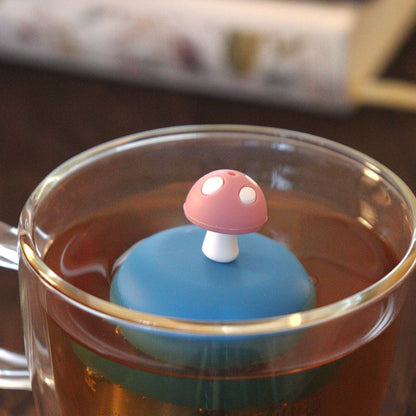 pink mushroom tea infuser for loose leaf tea on a blue base with a fine mesh stainless steel tea strainer that reads FLOAT2INFUSE floating in a glass tea mug with books in the background