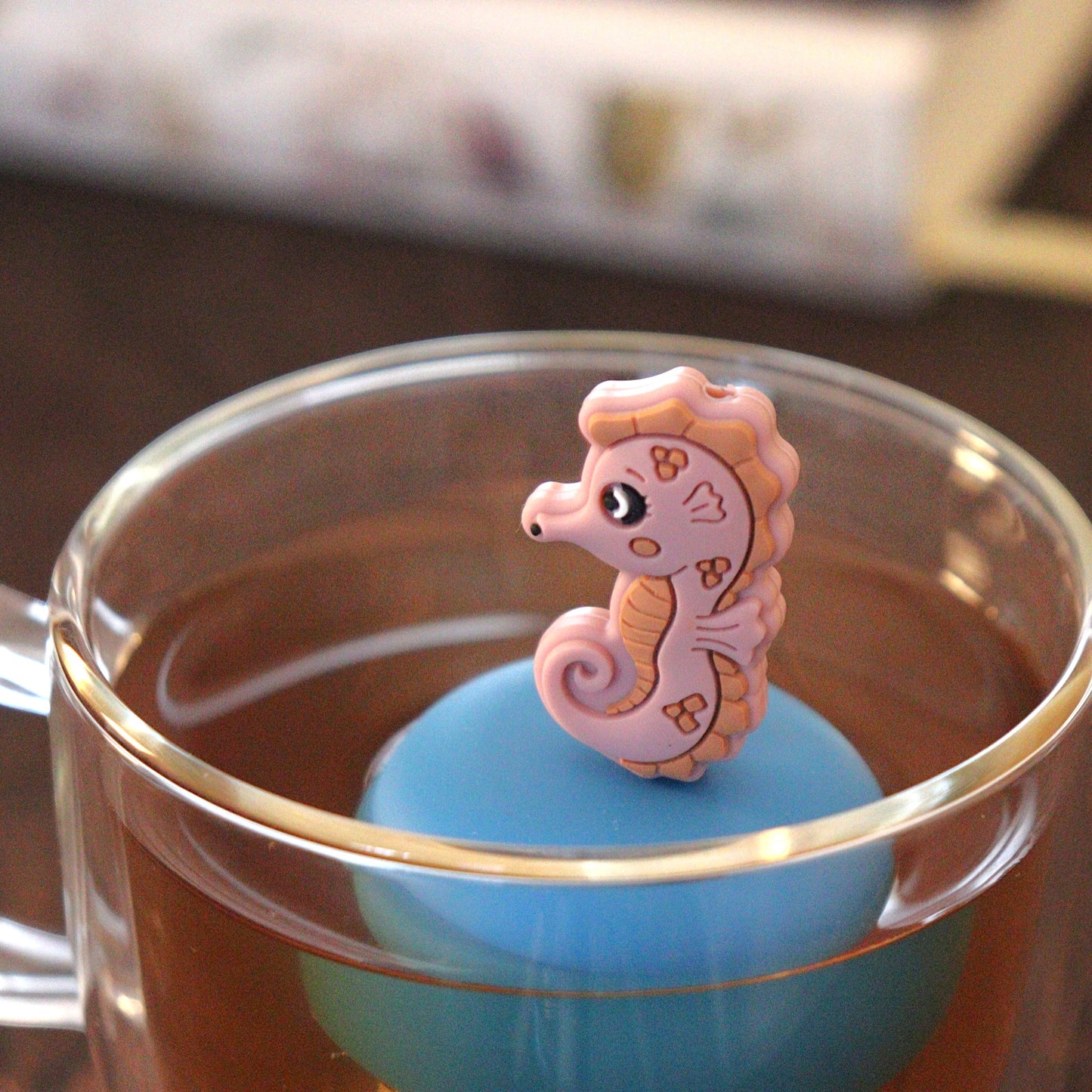 pink seahorse animal tea infuser for loose leaf tea  with a fine mesh stainless steel tea strainer that reads FLOAT2INFUSE on a blue base floating in a glass tea mug with books in the background