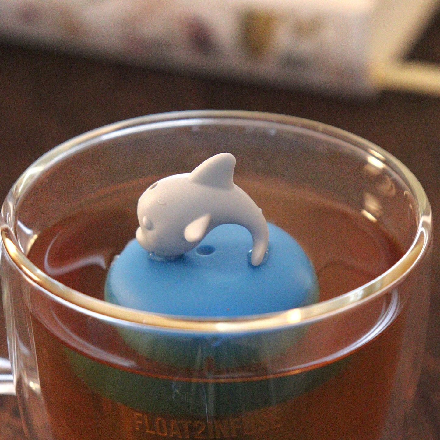 grey dolphin animal tea infuser for loose leaf tea with a fine mesh stainless steel tea strainer that reads FLOAT2INFUSE  on a blue base floating in a glass tea mug with books in the background