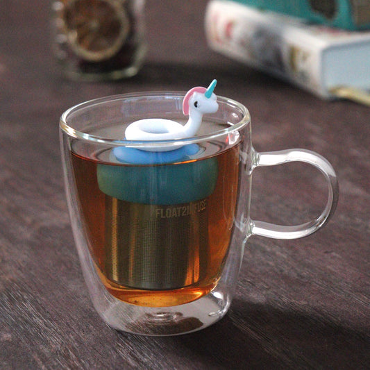 cute unicorn pool float tea infuser for loose leaf tea on a blue base with a fine mesh stainless steel tea strainer that reads FLOAT2INFUSE floating in a glass tea mug with loose leaf tea and books in the background