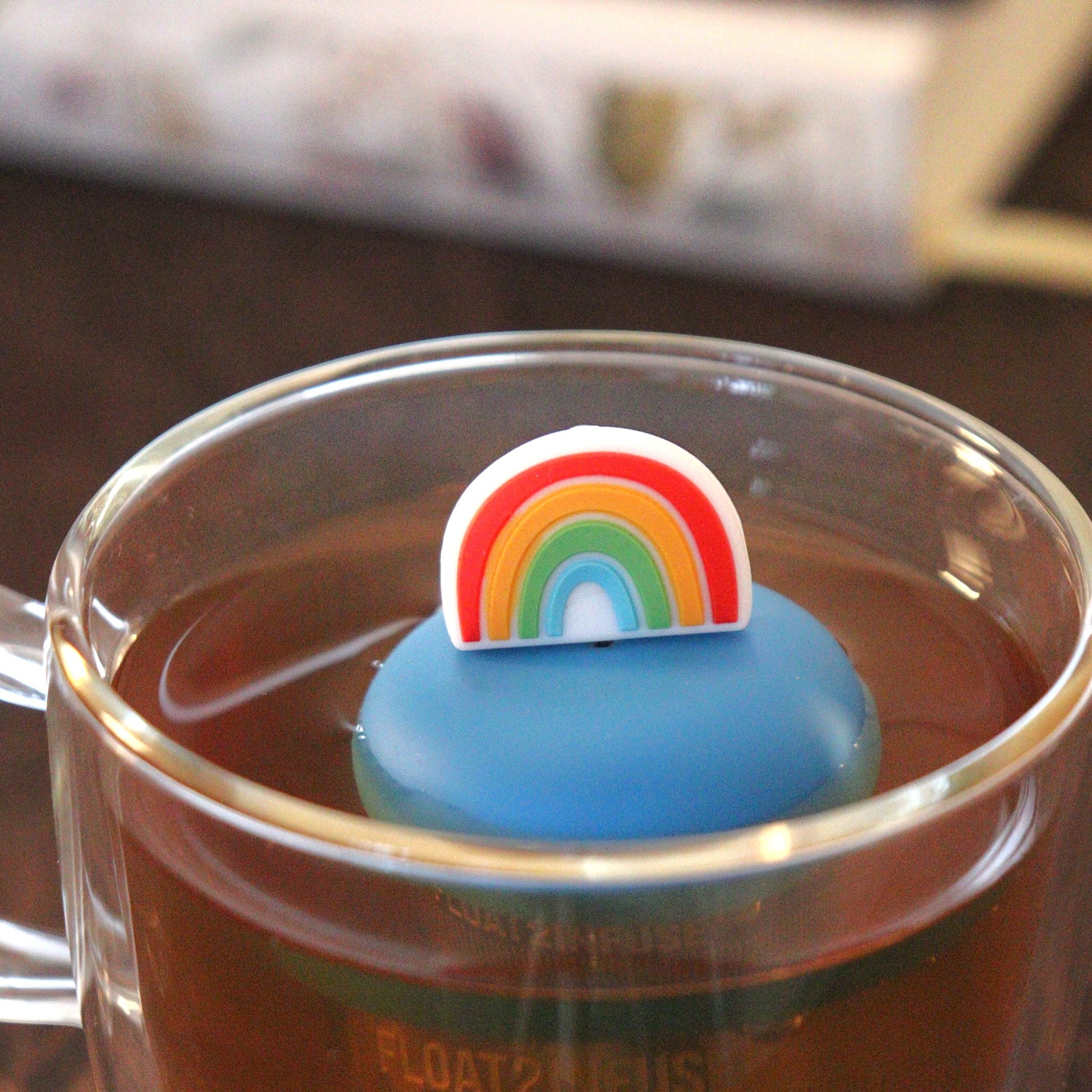 rainbow tea infuser for loose leaf tea with a fine mesh stainless steel tea strainer that reads FLOAT2INFUSE on a blue base floating in a glass tea mug with books in the background