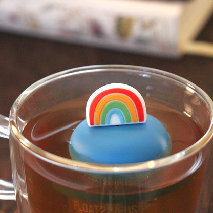 rainbow tea infuser for loose leaf tea with a fine mesh stainless steel tea strainer that reads FLOAT2INFUSE on a blue base floating in a glass tea mug with books in the background