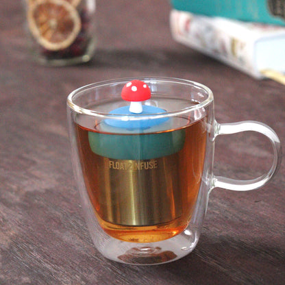 red mushroom tea infuser for loose leaf tea with a fine mesh stainless steel tea strainer that reads FLOAT2INFUSE on a blue base floating in a glass tea mug with loose leaf tea and books in the background