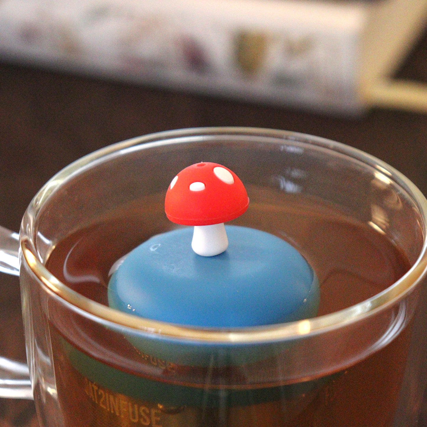 red mushroom tea infuser for loose leaf tea with a fine mesh stainless steel tea strainer that reads FLOAT2INFUSE on a blue base floating in a glass tea mug with books in the background