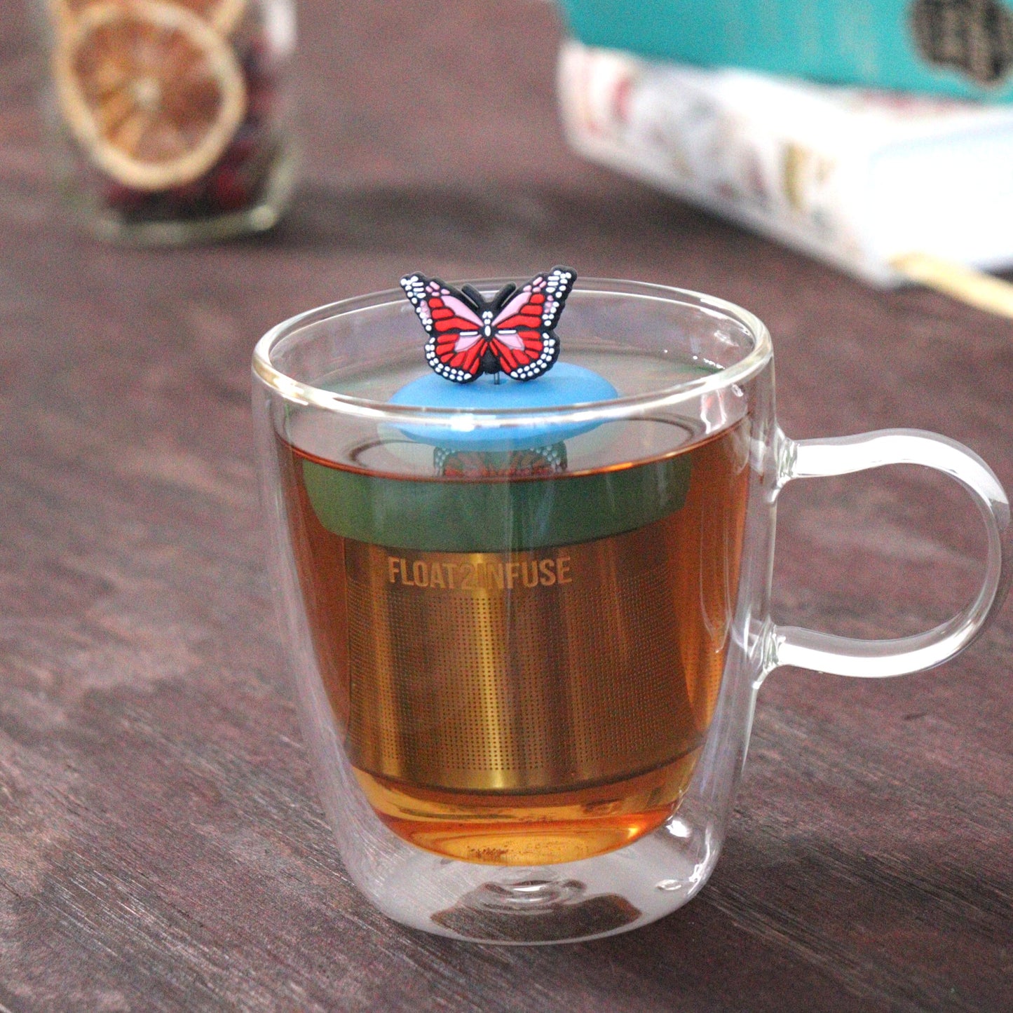 red and pink butterfly tea infuser for loose leaf tea  with a fine mesh stainless steel tea strainer that reads FLOAT2INFUSE on a blue base floating in a glass tea mug with loose leaf tea and books in the background