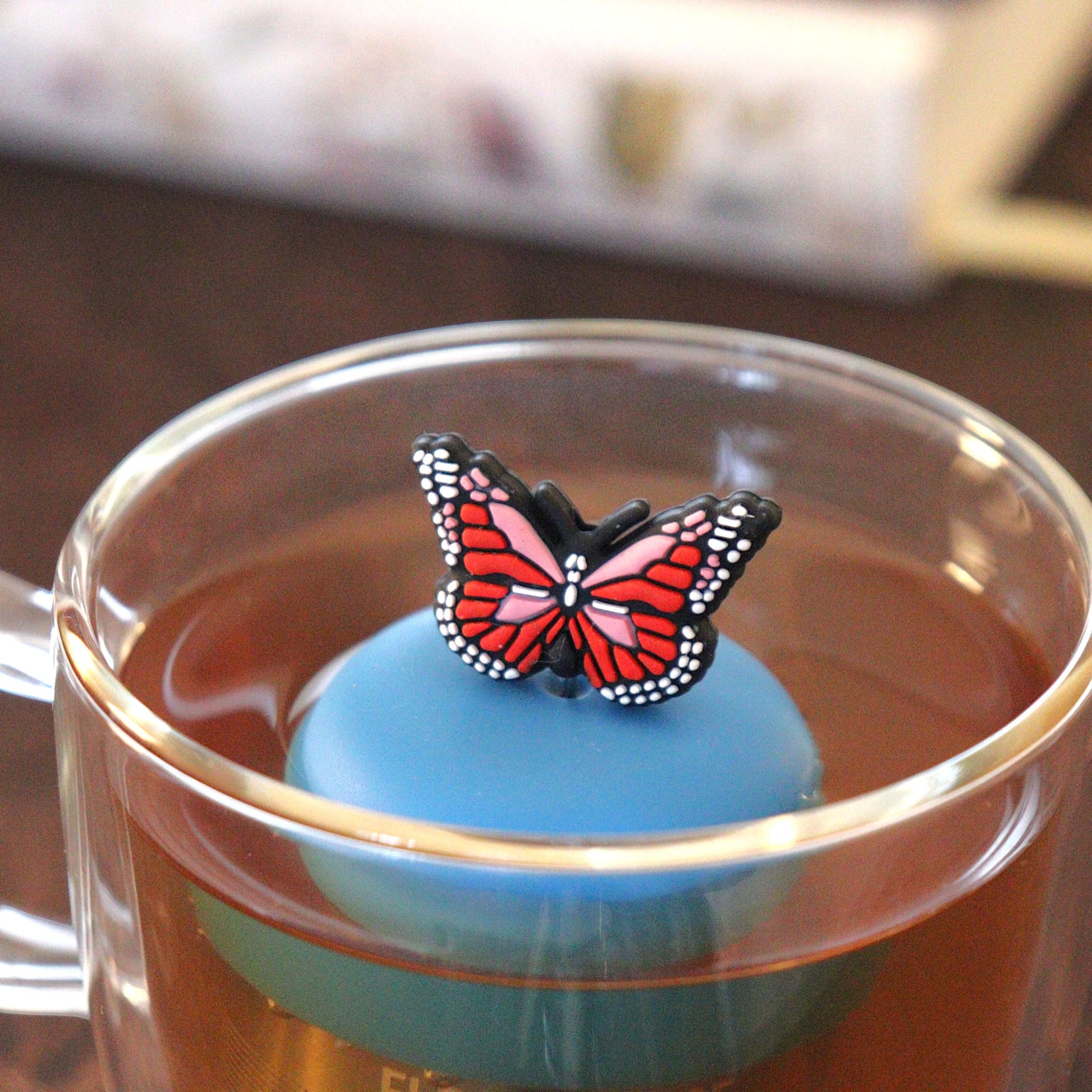 red and pink butterfly tea infuser for loose leaf tea  with a fine mesh stainless steel tea strainer that reads FLOAT2INFUSE on a blue base floating in a glass tea mug with books in the background