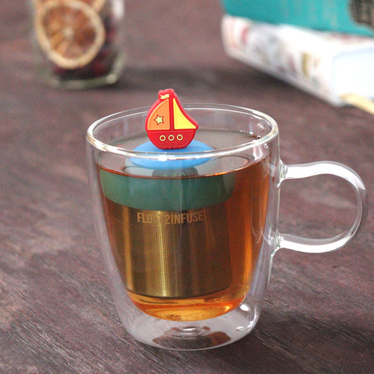red sailboat tea infuser for loose leaf tea  with a fine mesh stainless steel tea strainer that reads FLOAT2INFUSE on a blue base floating in a glass tea mug with loose leaf tea and books in the background