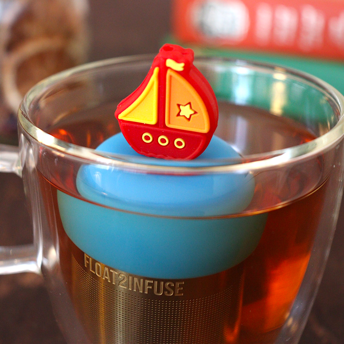 red sailboat tea infuser for loose leaf tea with a fine mesh stainless steel tea strainer that reads FLOAT2INFUSE on a blue base floating in a glass tea mug with loose leaf tea and books in the background