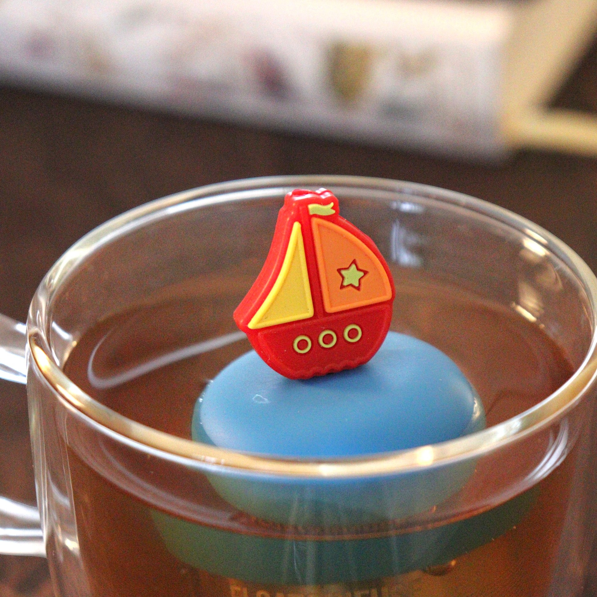 red sailboat tea infuser for loose leaf tea with a fine mesh stainless steel tea strainer that reads FLOAT2INFUSE on a blue base floating in a glass tea mug with books in the background
