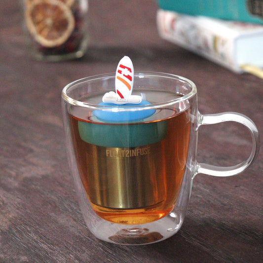 surf board tea infuser for loose leaf tea on a blue base with a fine mesh stainless steel tea strainer that reads FLOAT2INFUSE floating in a glass tea mug with loose leaf tea and books in the background