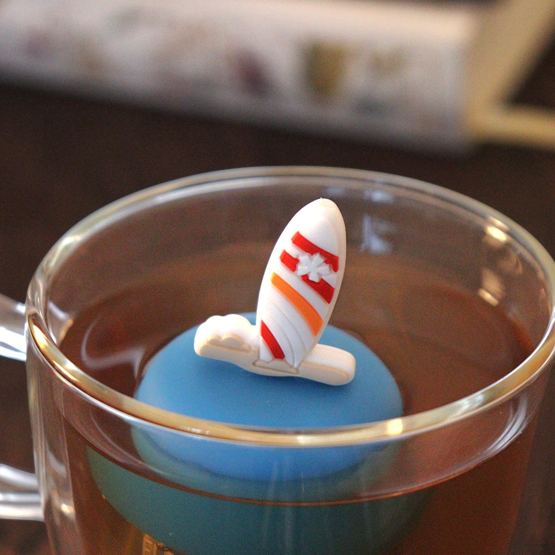 surf board tea infuser for loose leaf tea on a blue base with a fine mesh stainless steel tea strainer that reads FLOAT2INFUSE floating in a glass tea mug with books in the background