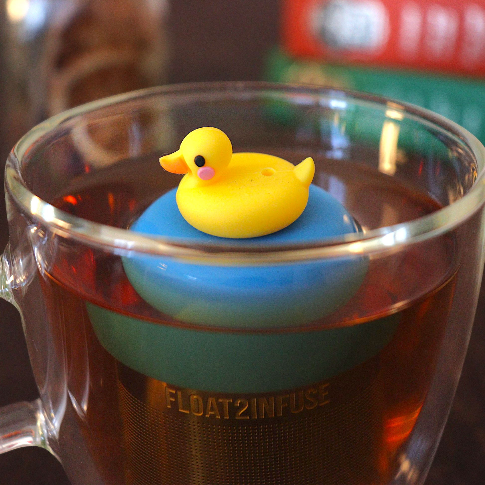 Yellow rubber duckie tea infuser for loose leaf tea with a fine mesh stainless steel tea strainer 
that reads FLOAT2INFUSE on a blue base with a book and loost leaf tea in the background