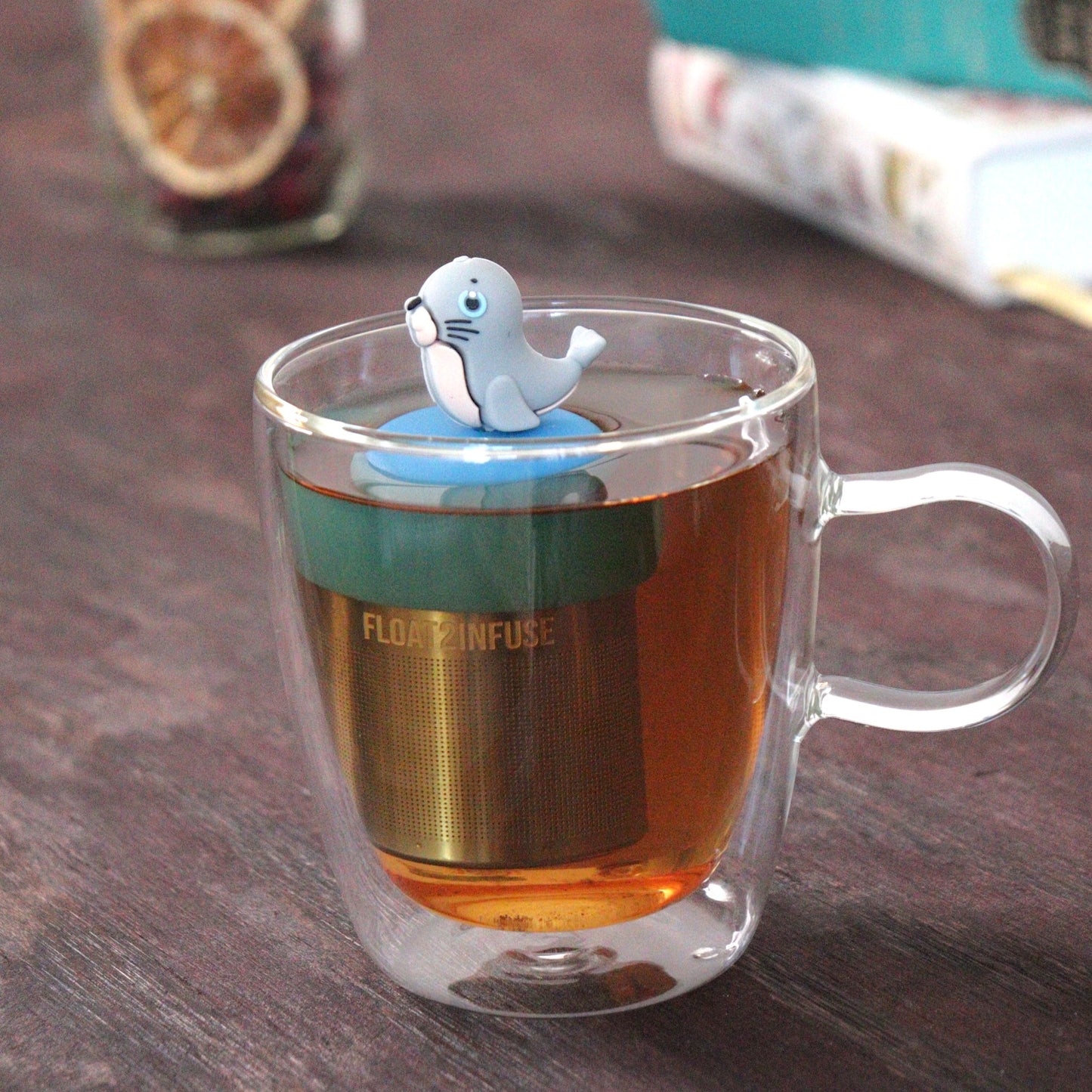 cute grey seal animal tea infuser for loose leaf tea  with a fine mesh stainless steel tea strainer that reads FLOAT2INFUSE on a blue base floating in a glass tea mug with loose leaf tea and books in the background