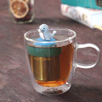cute grey seal animal tea infuser for loose leaf tea  with a fine mesh stainless steel tea strainer that reads FLOAT2INFUSE on a blue base floating in a glass tea mug with loose leaf tea and books in the background
