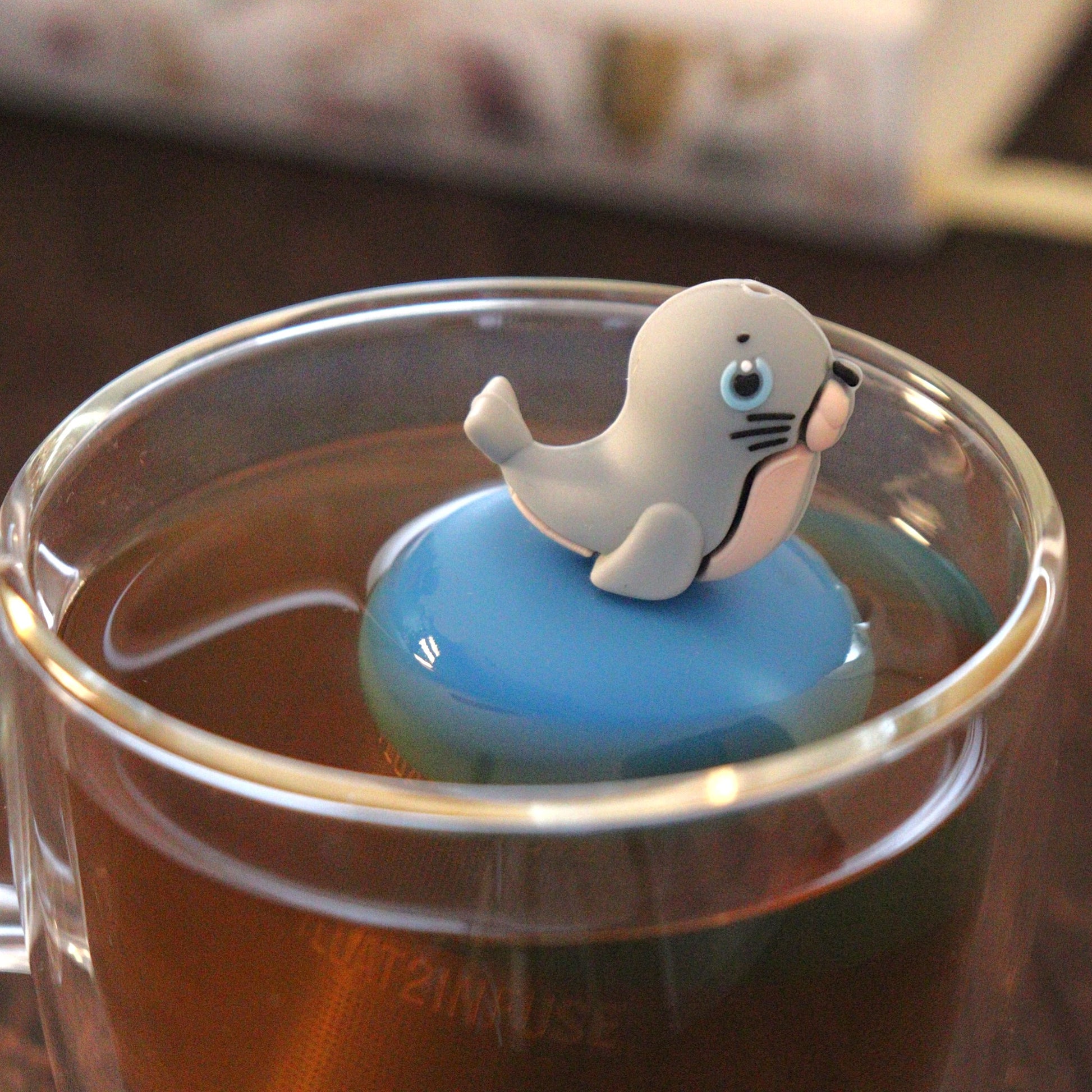 cute grey seal animal tea infuser for loose leaf tea  with a fine mesh stainless steel tea strainer that reads FLOAT2INFUSE on a blue base floating in a glass tea mug with books in the background