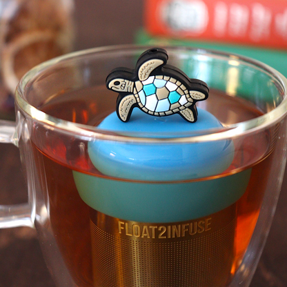 A sea turtle with a geometric shell pattern standing on its fins tea infuser for loose leaf tea with a fine mesh stainless steel tea strainer 
that reads FLOAT2INFUSE on a blue base with loose leaf tea and books in the background
