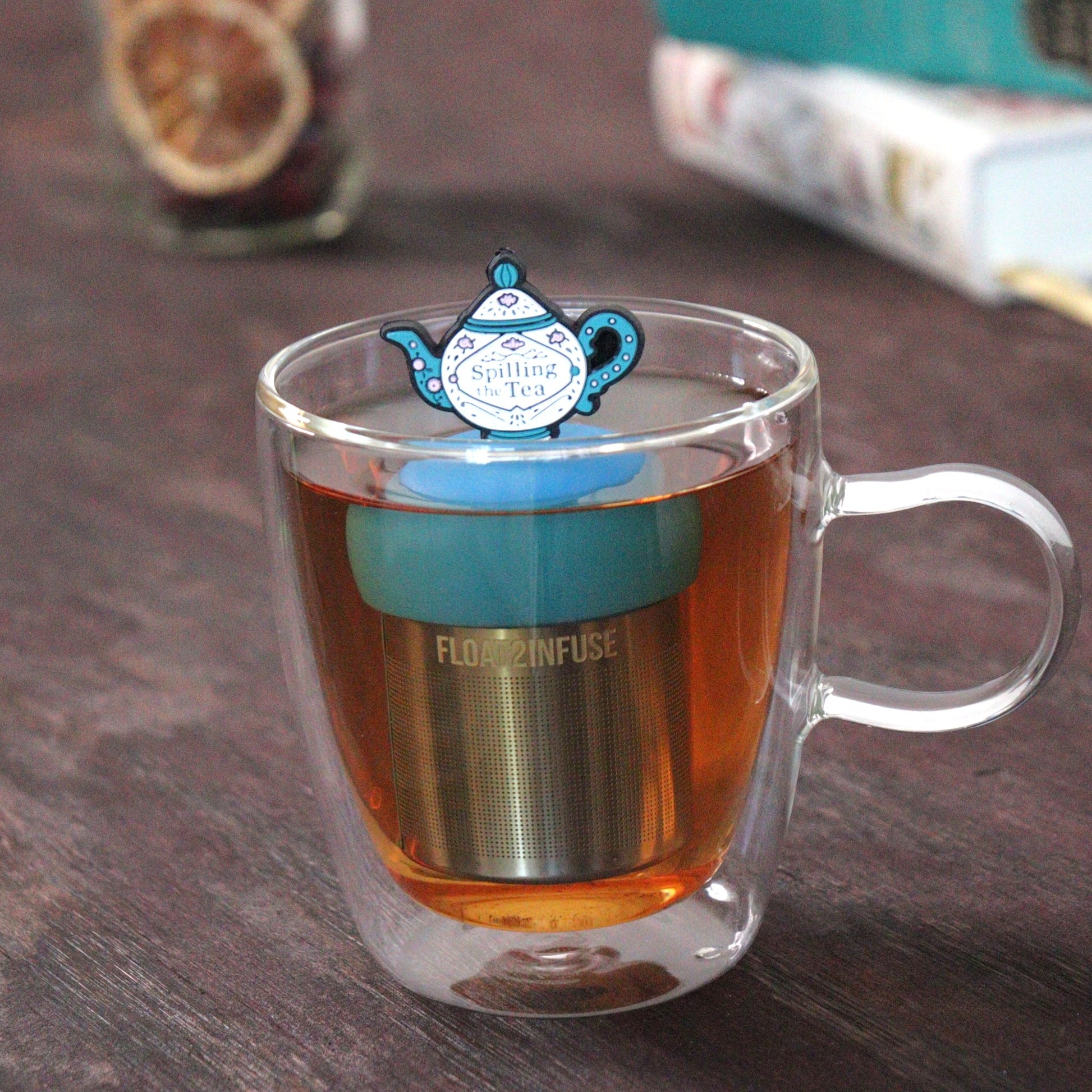 blue and white teapot that reads "spill the  tea" tea infuser for loose leaf tea  with a fine mesh stainless steel tea strainer that reads FLOAT2INFUSE on a blue base floating in a glass tea mug with loose leaf tea and  books in the background