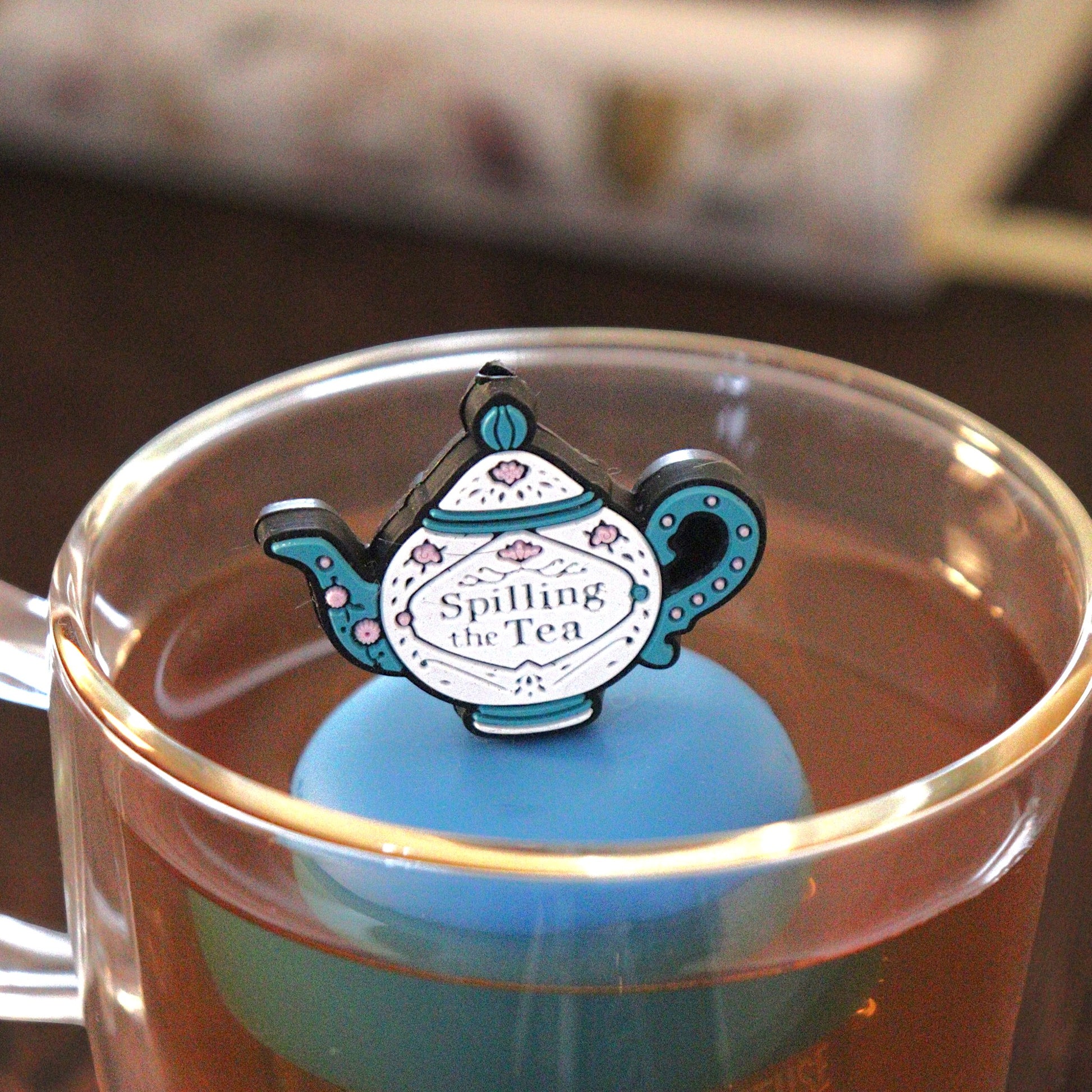 blue and white teapot that reads "spill the  tea" tea infuser for loose leaf tea  with a fine mesh stainless steel tea strainer that reads FLOAT2INFUSE on a blue base floating in a glass tea mug with books in the background