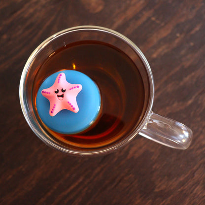 looking down on a pink starfish animal tea infuser for loose leaf tea  with a fine mesh stainless steel tea strainer that reads FLOAT2INFUSE on a blue basefloating in a glass tea mug