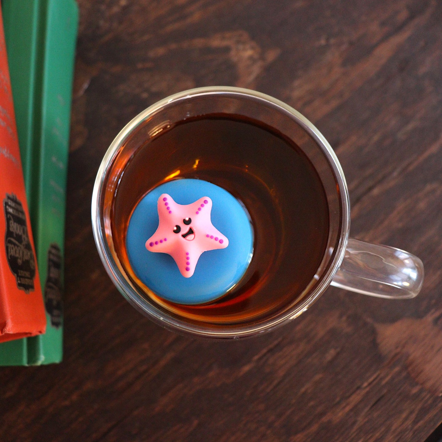 looking down on a pink starfish animal tea infuser for loose leaf tea  with a fine mesh stainless steel tea strainer that reads FLOAT2INFUSE on a blue base floating in a glass tea mug