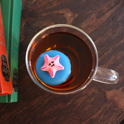 looking down on a pink starfish animal tea infuser for loose leaf tea  with a fine mesh stainless steel tea strainer that reads FLOAT2INFUSE on a blue base floating in a glass tea mug