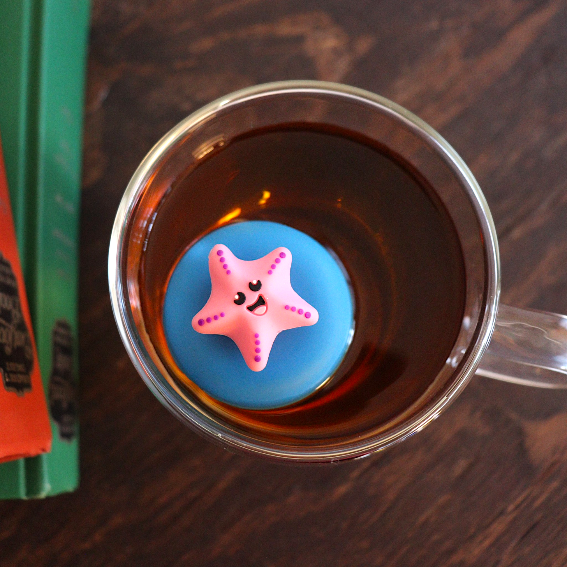 looking down on a pink starfish animal tea infuser for loose leaf tea  with a fine mesh stainless steel tea strainer that reads FLOAT2INFUSE on a blue base floating in a glass tea mug