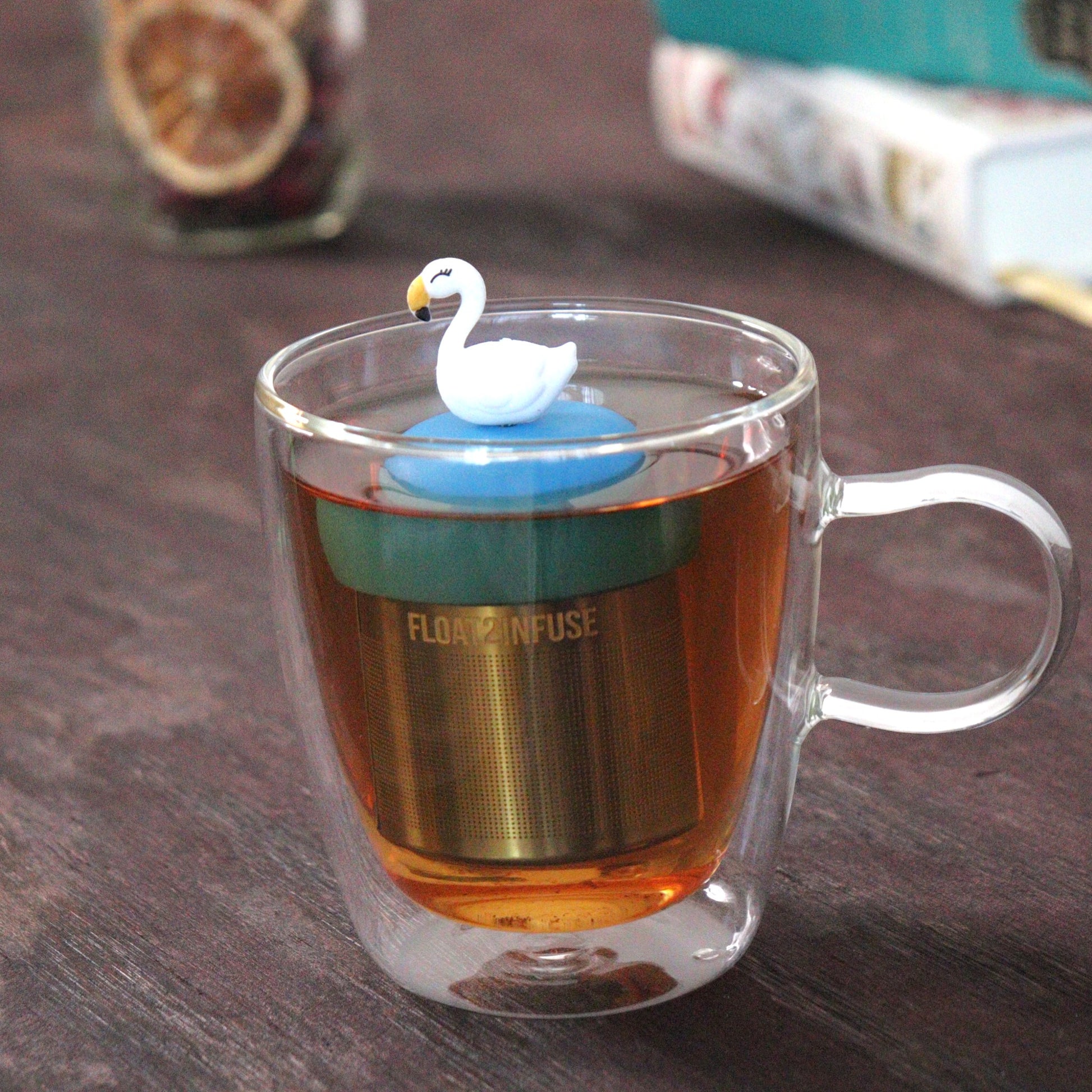 white swan animal tea infuser for loose leaf tea with a fine mesh stainless steel tea strainer that reads FLOAT2INFUSE on a blue base floating in a glass tea mug with loose leaf tea and books in the background