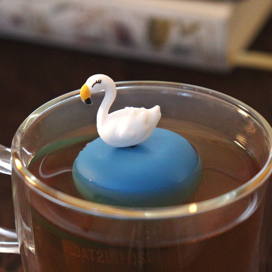 white swan animal tea infuser for loose leaf tea with a fine mesh stainless steel tea strainer that reads FLOAT2INFUSE on a blue base  floating in a glass tea mug with books in the background
