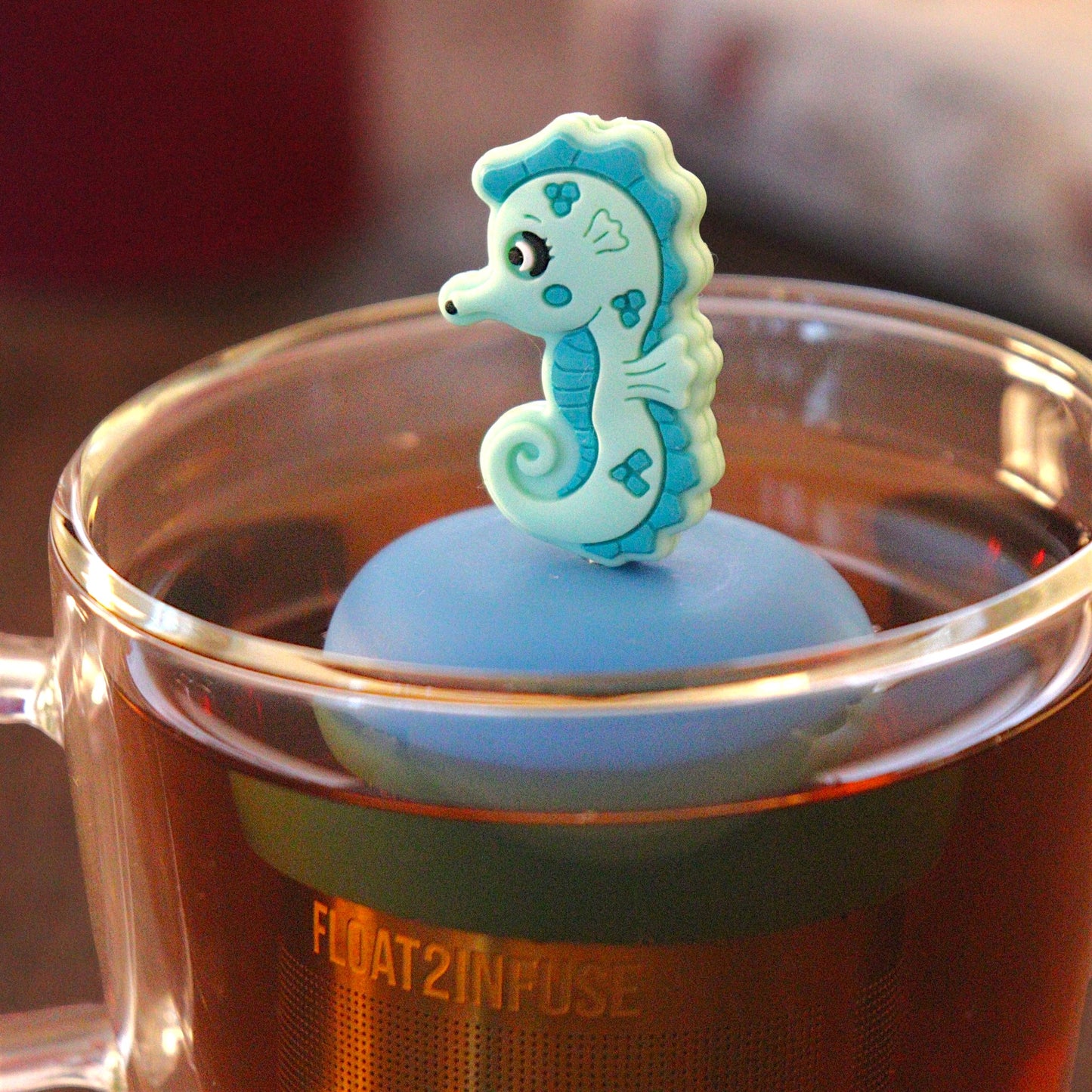 teal seahorse  tea infuser for loose leaf tea with a fine mesh stainless steel tea infuser that reads FLOAT2INFUSE on a blue base  floating in a glass tea mug with  loose leaf tea and books in the background