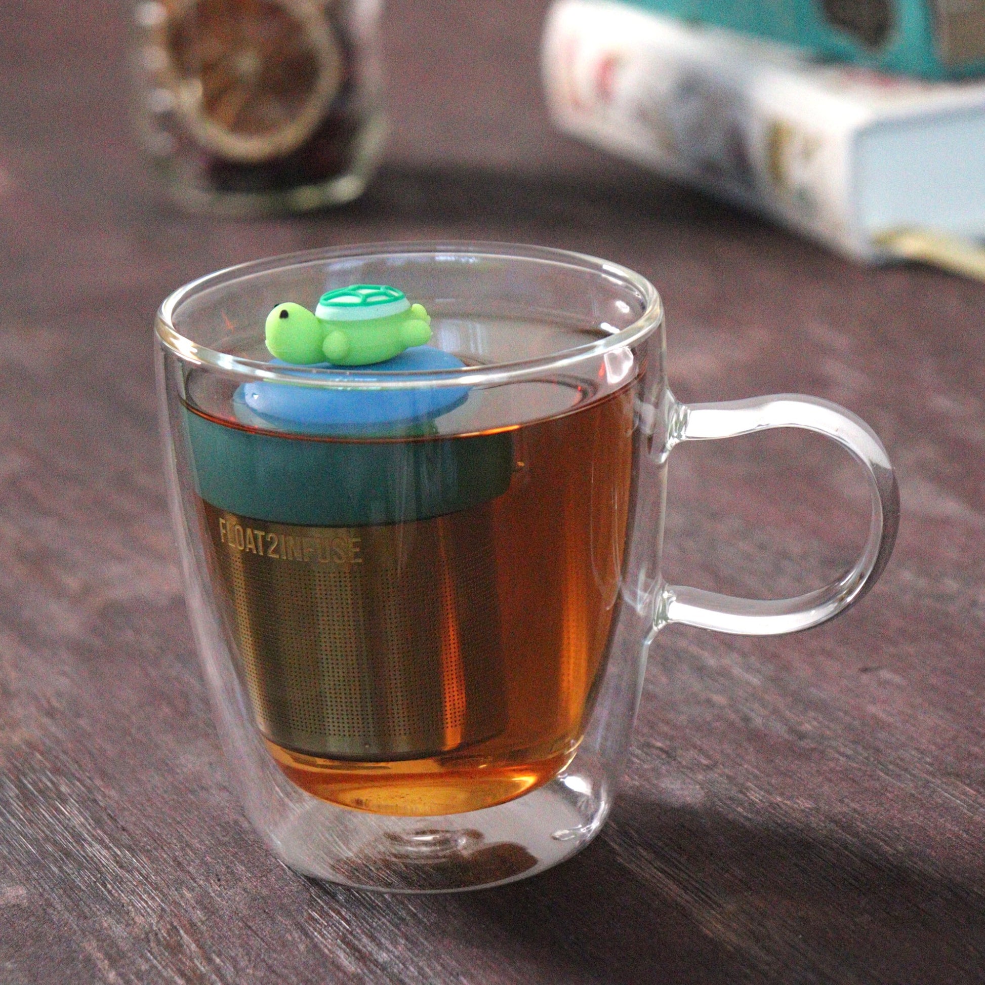 green turtle animal tea infuser for loose leaf tea with a fine mesh stainless steel tea strainer that reads FLOAT2INFUSE on a blue base floating in a glass tea mug with loose leaf tea and books in the background
