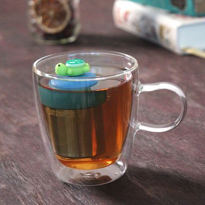 green turtle animal tea infuser for loose leaf tea with a fine mesh stainless steel tea strainer that reads FLOAT2INFUSE on a blue base floating in a glass tea mug with loose leaf tea and books in the background