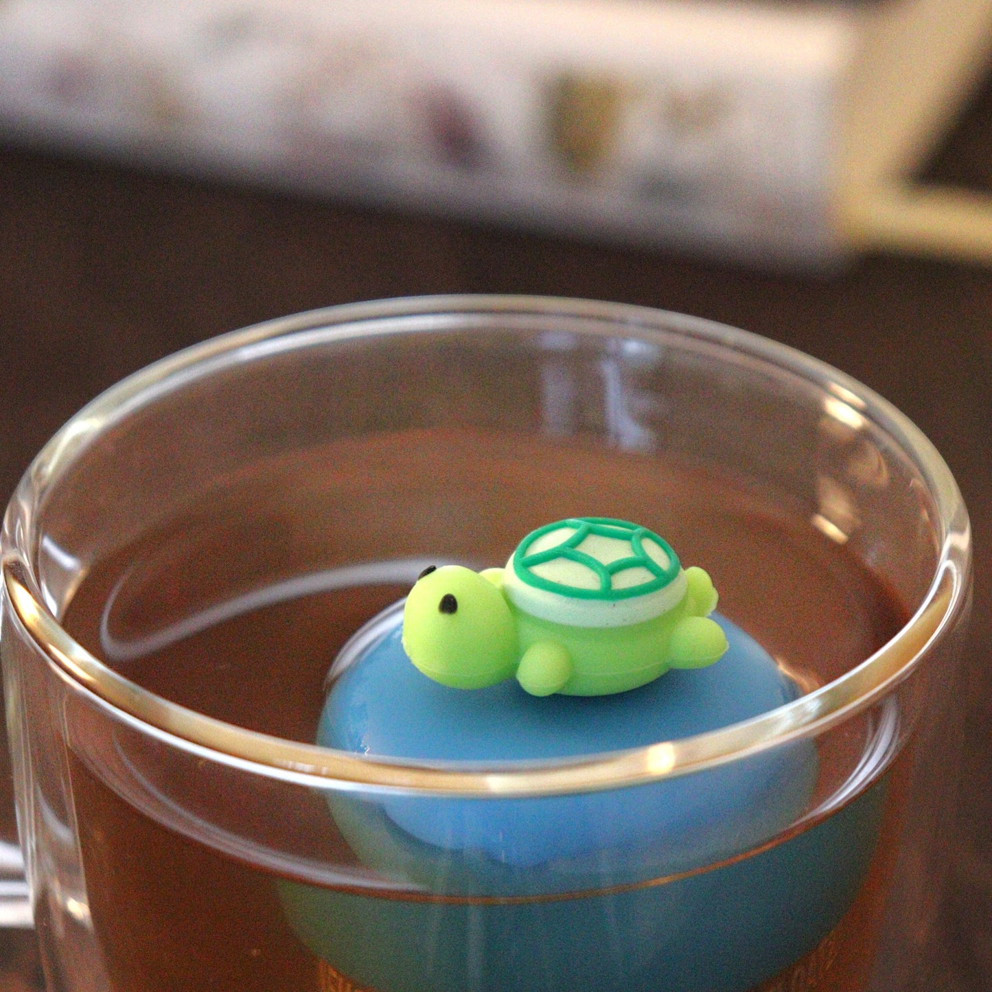 green turtle animal tea infuser for loose leaf tea on a blue base with a fine mesh stainless steel tea strainer that reads FLOAT2INFUSE floating in a glass tea mug with books in the background