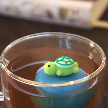 green turtle animal tea infuser for loose leaf tea on a blue base with a fine mesh stainless steel tea strainer that reads FLOAT2INFUSE floating in a glass tea mug with books in the background