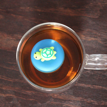 top view of a green turtle animal tea infuser for loose leaf tea with a fine mesh stainless steel tea strainer that reads FLOAT2INFUSE on a blue base floating in a glass tea mug