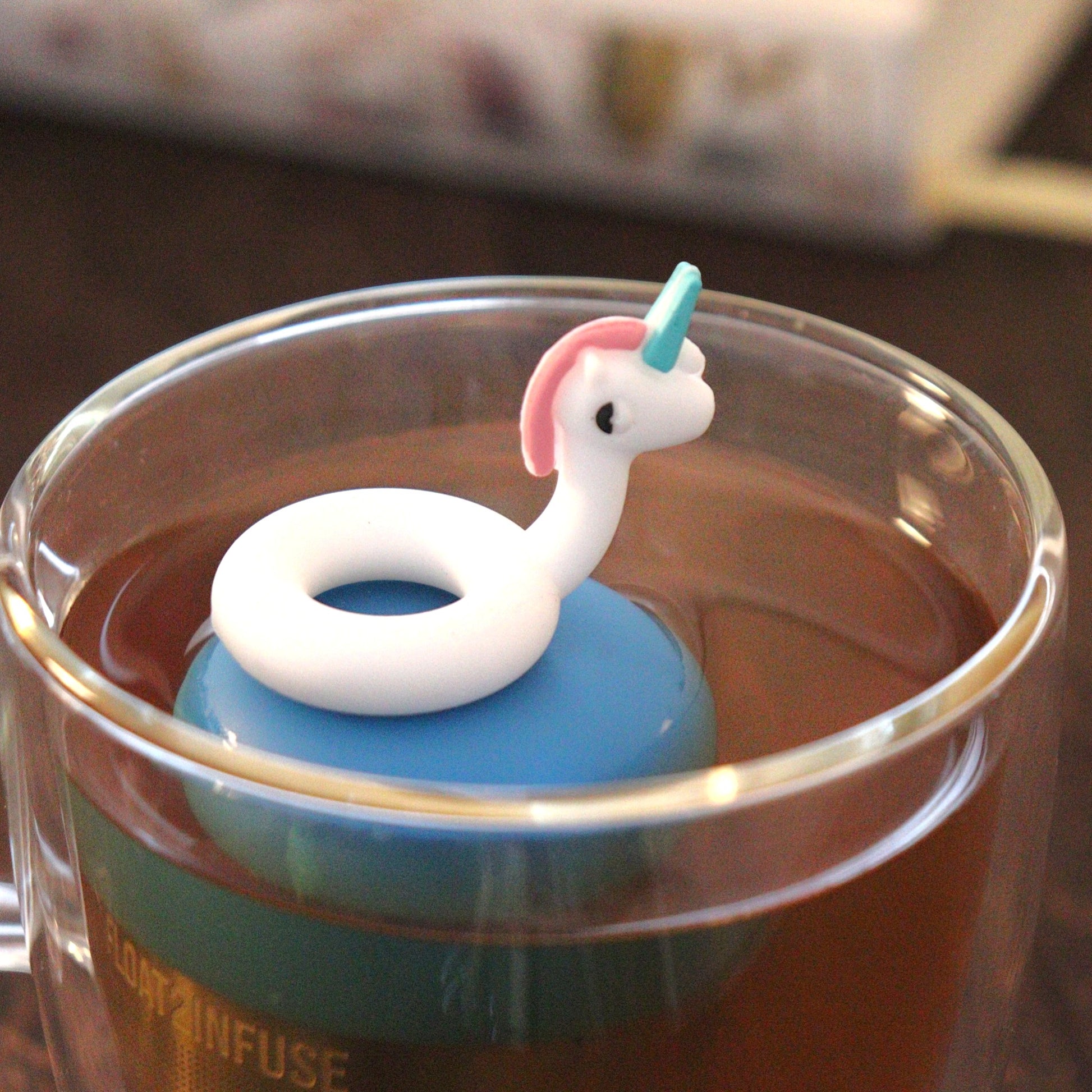 unicorn pool float tea infuser for loose leaf tea on a blue base with a fine mesh stainless steel tea strainer that reads FLOAT2INFUSE floating in a glass tea mug with books in the background