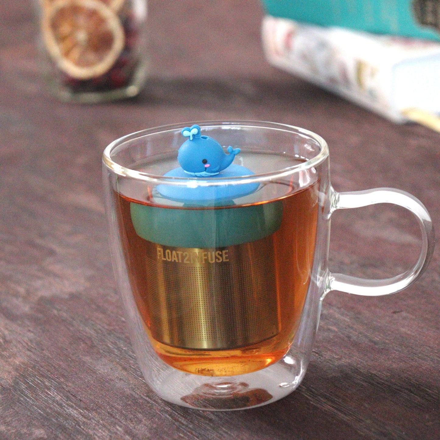 blue water whale animal tea infuser for loose leaf tea  with a fine mesh stainless steel tea strainer that reads FLOAT2INFUSE on a blue base floating in a glass tea mug with loose leaf tea and books in the background