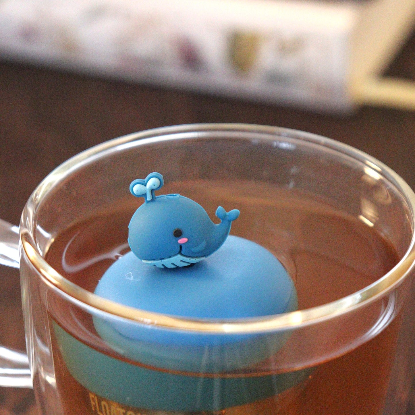blue water whale animal tea infuser for loose leaf tea  with a fine mesh stainless steel tea strainer that reads FLOAT2INFUSE on a blue base floating in a glass tea mug with books in the background