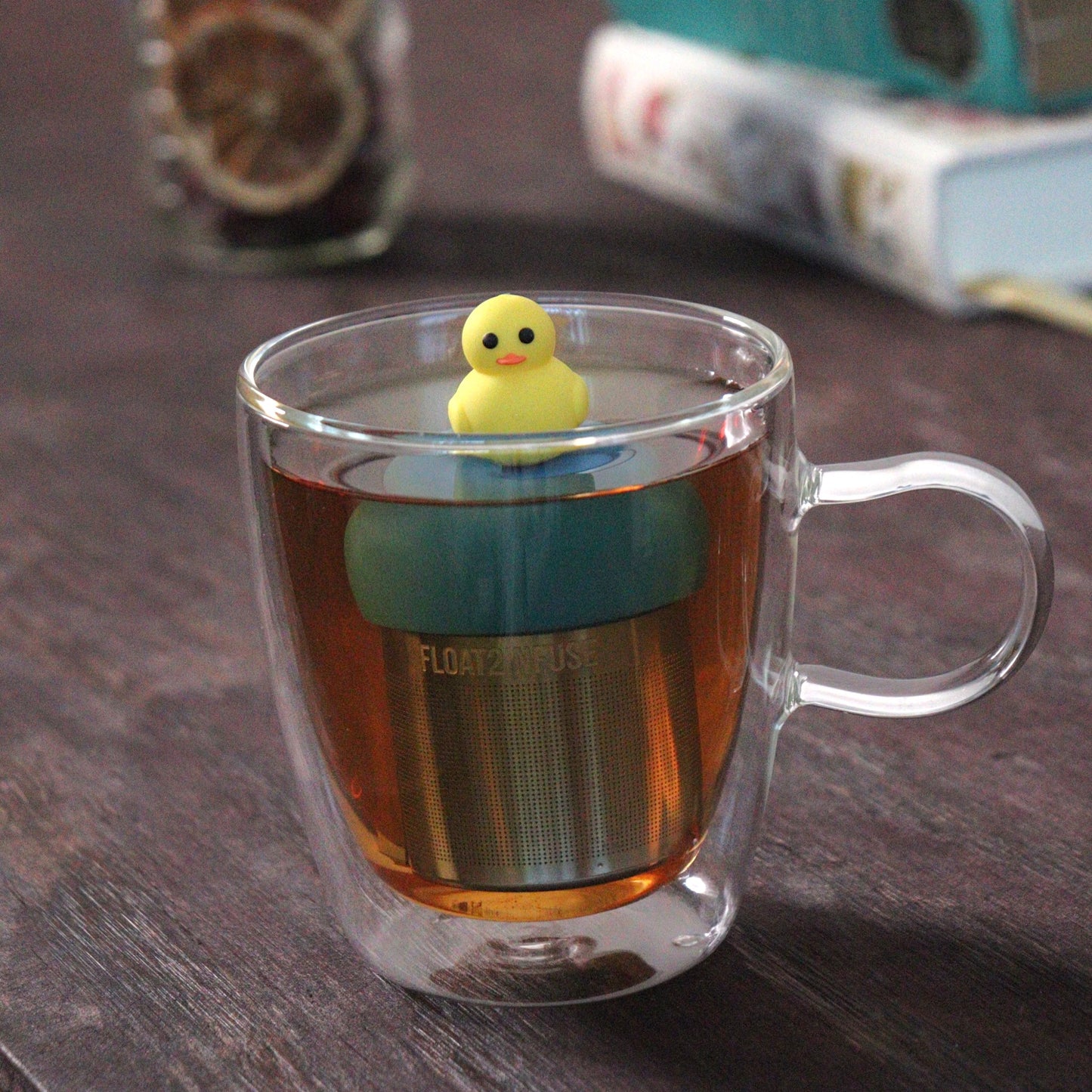 yellow duck animal tea infuser for loose leaf tea  with a fine mesh stainless steel tea strainer that reads FLOAT2INFUSE on a blue base floating in a glass tea mug with loose leaf tea and books in the background