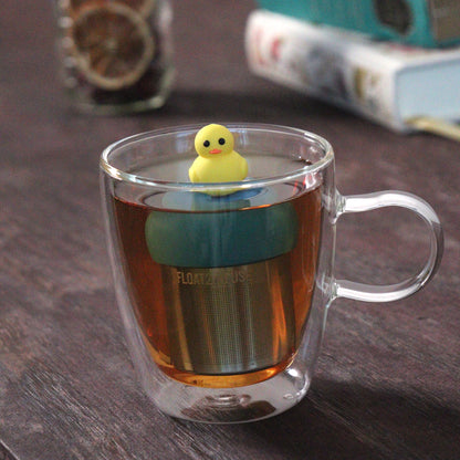 yellow duck animal tea infuser for loose leaf tea  with a fine mesh stainless steel tea strainer that reads FLOAT2INFUSE on a blue base floating in a glass tea mug with loose leaf tea and books in the background