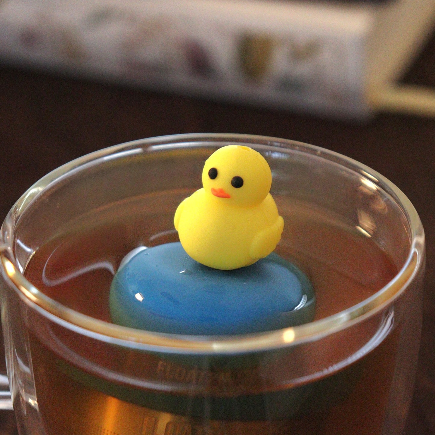 yellow duck animal tea infuser for loose leaf tea  with a fine mesh stainless steel tea strainer that reads FLOAT2INFUSE on a blue base floating in a glass tea mug with books in the background