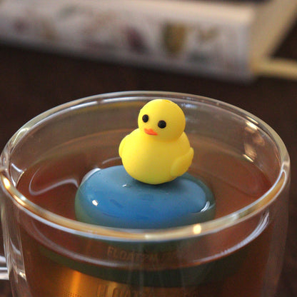 yellow duck animal tea infuser for loose leaf tea  with a fine mesh stainless steel tea strainer that reads FLOAT2INFUSE on a blue base floating in a glass tea mug with books in the background