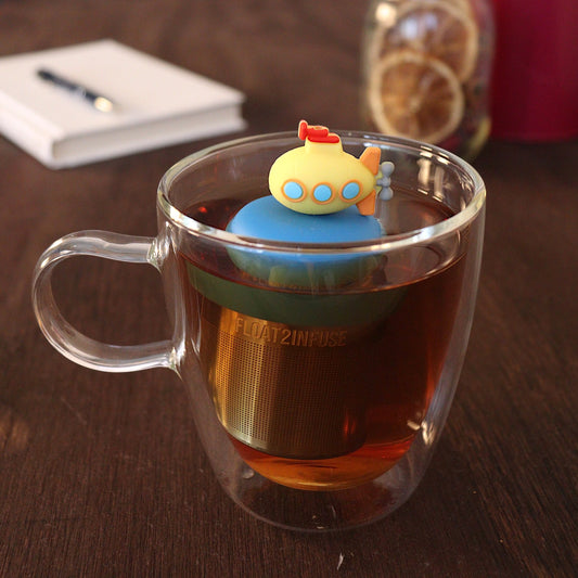 Cute yellow submarine tea infuser for loose leaf tea with a fine mesh stainless steel tea strainer 
that reads FLOAT2INFUSE on a blue base with a pen laying across a book and dried orange slices in a mason jar in the background