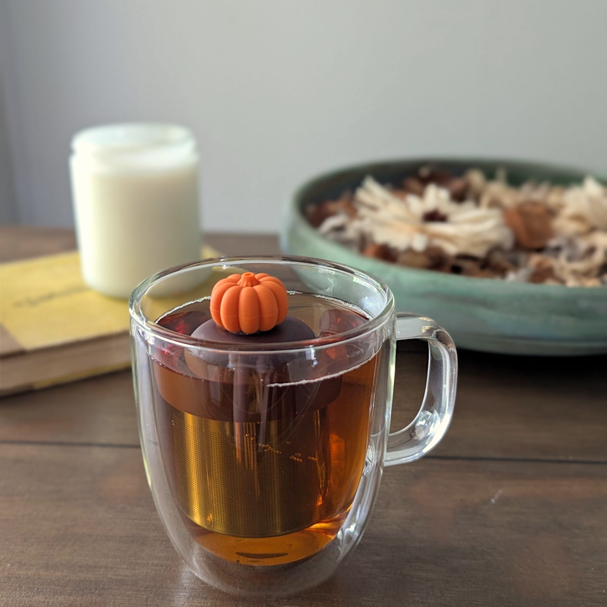 3d orange pumpkin tea infuser for loose leaf tea with fine mesh stainless steel tea strainer that reads FLOAT2INFUSE on a brown base floating in a glass tea mug with a candle and potpourri in the background