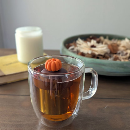 3d orange pumpkin tea infuser for loose leaf tea with fine mesh stainless steel tea strainer that reads FLOAT2INFUSE on a brown base floating in a glass tea mug with a candle and potpourri in the background