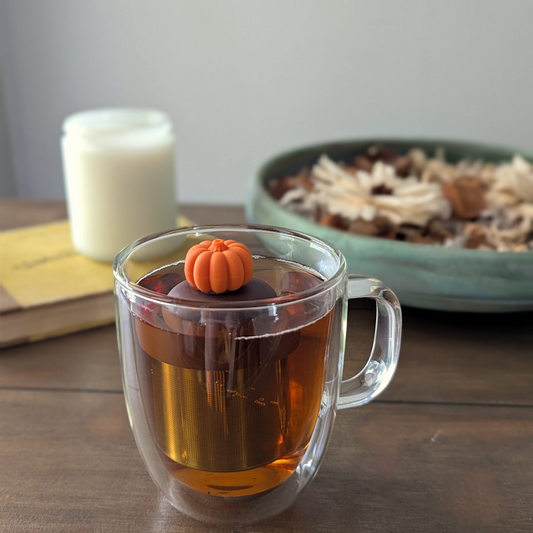 3d orange pumpkin tea infuser for loose leaf tea with fine mesh stainless steel tea strainer that reads FLOAT2INFUSE on a brown base floating in a glass tea mug with a candle and potpourri in the background