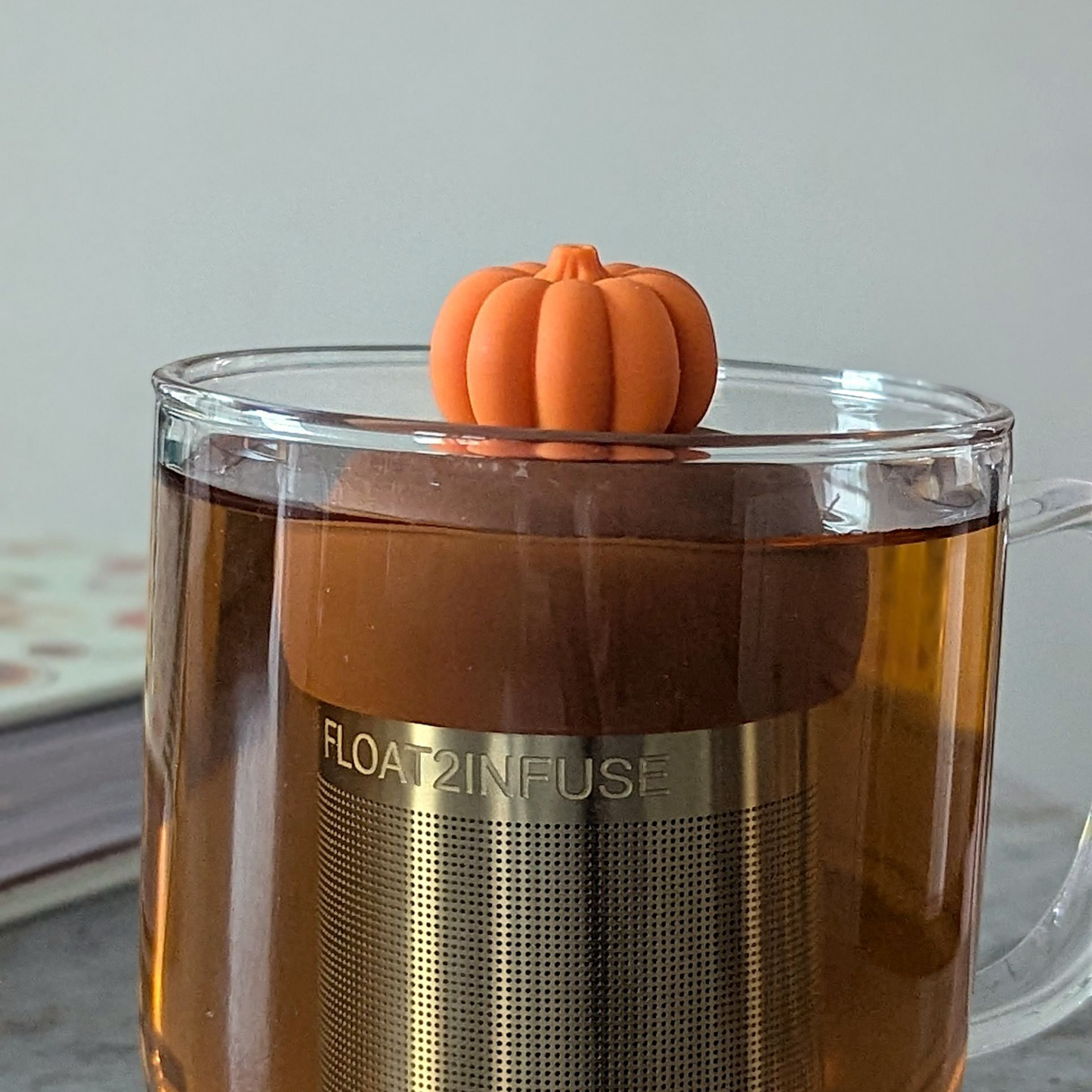 3d orange pumpkin tea infuser for loose leaf tea with fine mesh stainless steel tea strainer that reads FLOAT2INFUSE on a brown base floating in a glass tea mug with a book in the background