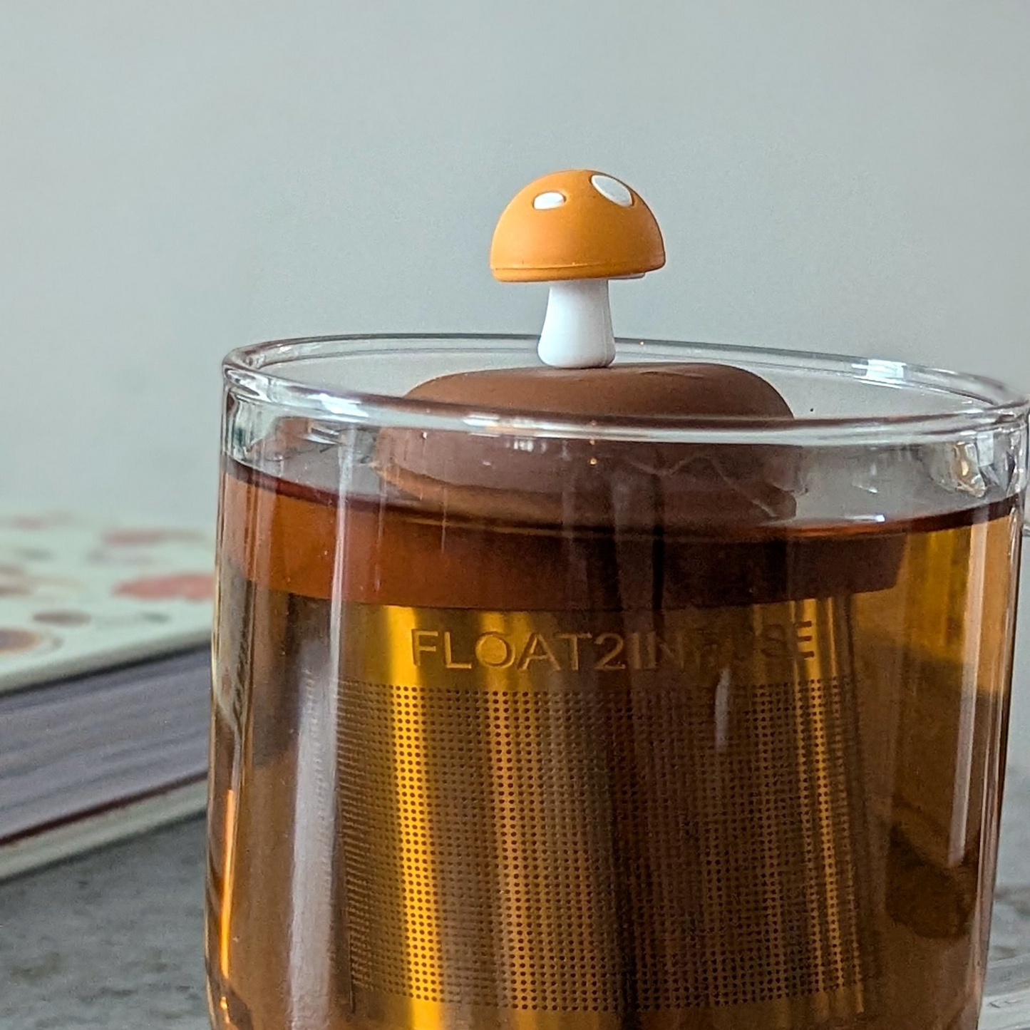 amber yellow mushroom with white dots tea infuser for loose leaf tea with a fine mesh stainless steel tea strainer that reads FLOAT2INFUSE on a brown base floating in a glass tea mug with a book in the background