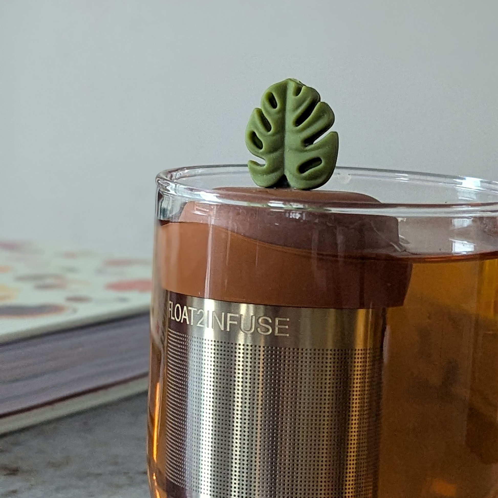 army green monstera leaf tea infuser for loose leaf tea  with a fine mesh stainless steel tea steeper that reads FLOAT2INFUSE on a brown base floating in a glass tea mug with books in the background